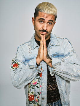 Young trendy guy in stylish denim jacket begging with folded hands near chin and looking at camera against gray background