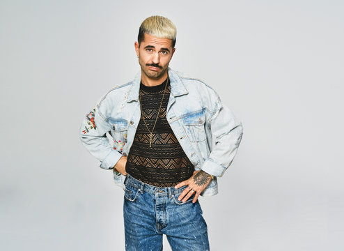 Young ethnic man making grimace doubting face looking at camera wearing trendy denim jacket with floral pattern while standing against gray background