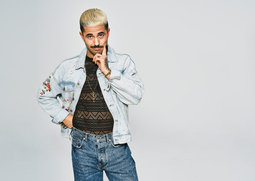 Young ethnic man making grimace doubting face with finger looking at camera wearing trendy denim jacket with floral pattern while standing against gray background