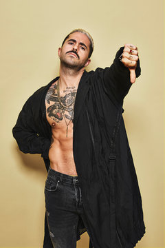 Young provocative male in black coat over naked tattooed torso showing thumbs down gesture in disapproval while standing against beige background