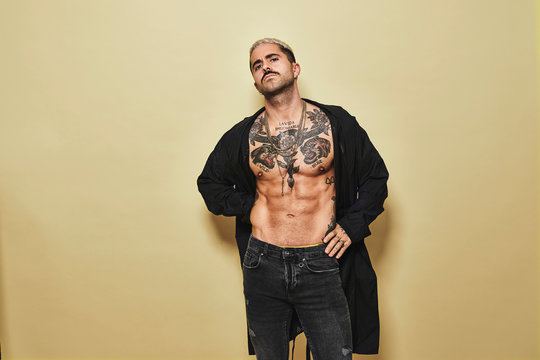 Confident arrogant stylish man with muscular tattooed torso wearing black coat and jeans looking at camera against beige background