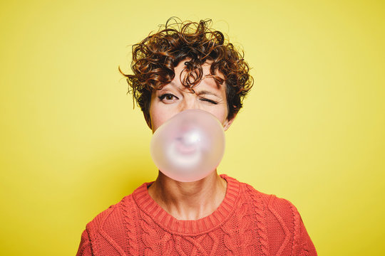 Young curly haired female in orange sweater blowing bubble gum and blinking looking to the camera while standing against yellow background