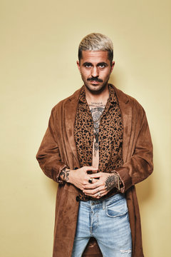Fashionable male model with tattoos wearing trendy coat over leopard shirt and jeans standing against beige background and looking at camera