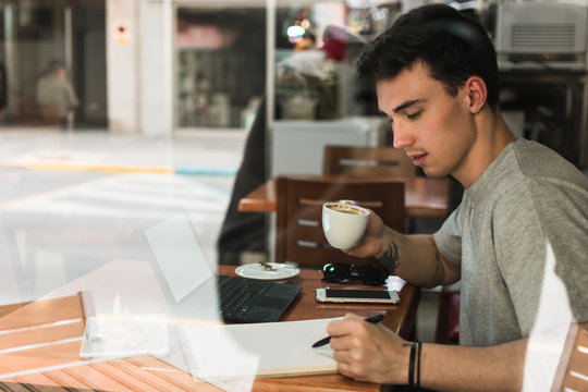 Side view of young man with cup of coffee writing in notepad while sitting at table behind glass and studying in cafeteria