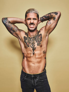 Handsome sexy attractive muscular male with various tattoos on naked torso and arms looking at camera making faces with mouth while standing against beige background