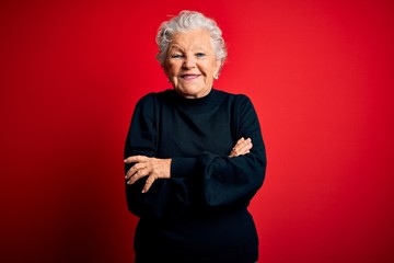 Senior beautiful woman wearing casual sweater standing over isolated red background happy face smiling with crossed arms looking at the camera. Positive person.