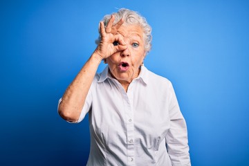 Senior beautiful woman wearing elegant shirt standing over isolated blue background doing ok gesture shocked with surprised face, eye looking through fingers. Unbelieving expression.