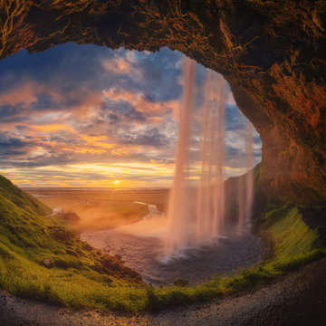 Majestic waterfall located on entrance of grassy stone cave against cloudy sundown sky on seashore in Iceland