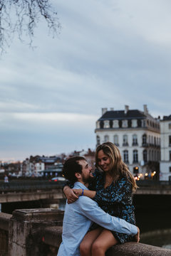 Happy young female sitting on old stone fence and embracing affectionate boyfriend while spending summer evening together in Bayonne