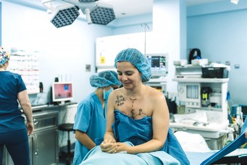 Adult woman with marks on breast and chest sitting on operating table near surgical team before plastic surgery in modern clinic