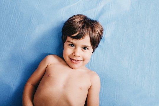 From above of cheerful shirtless little boy looking at camera while lying on blue hospital bed