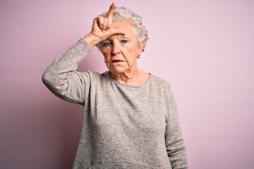 Senior beautiful woman wearing casual t-shirt standing over isolated pink background making fun of people with fingers on forehead doing loser gesture mocking and insulting.