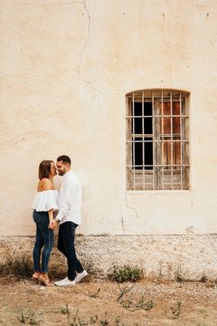 Full body happy man and woman holding hands and touching noses while standing near aged cottage with grated window