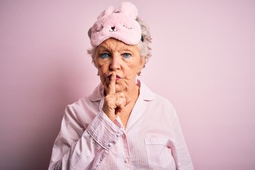 Senior beautiful woman wearing sleep mask and pajama over isolated pink background asking to be quiet with finger on lips. Silence and secret concept.