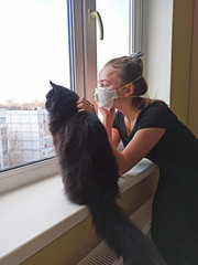 girl in protective mask with cat looking sadly out the window during quarantine