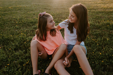 High angle of cheerful teen girl hugging little sister and looking at camera while sitting together on grassy meadow and enjoying sunny summer day in nature