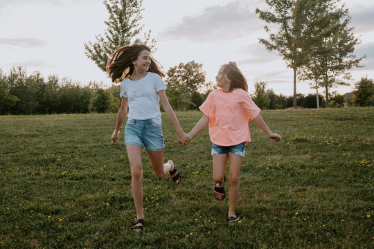 Cheerful carefree teen girl and youngest sister holding hands and running together on green meadow in summer day in countryside