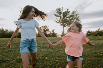 Cheerful carefree teen girl and youngest sister holding hands and running together on green meadow in summer day in countryside
