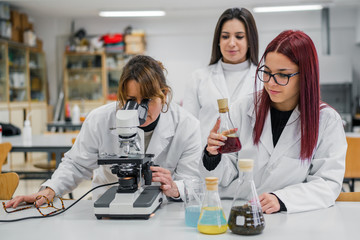 Adult woman examining sample in microscope near young women with chemical liquids during university lesson in modern lab