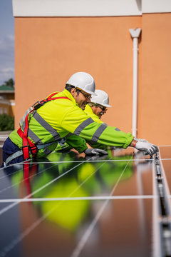Group of workers in uniform and helmets installing photovoltaic panels on roof of wooden construction near house