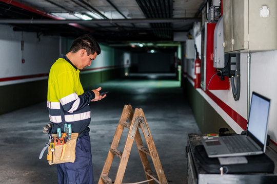 Side view of skilled man engineer in uniform using mobile phone while examining electrical equipment in modern building