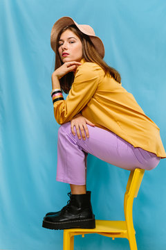 Side view of trendy confident millennial female in colorful clothes and hat sitting on stool and looking at camera against blue background