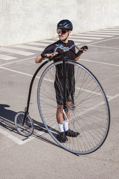 From above pensive man in black tracksuit and helmet looking away while standing in parking lot with retro high wheel bicycle against concrete wall in sunny day