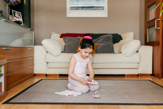 Cute girl in leotard and tights sitting on floor near sofa and putting on dance shoes before ballet rehearsal at home