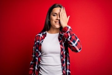 Young beautiful brunette woman wearing casual shirt standing over isolated red background covering one eye with hand, confident smile on face and surprise emotion.