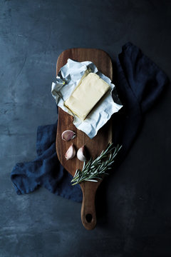 Top view of opened pack of butter in composition with cloves of garlic and rosemary stems on wooden cutting board and textile placed on dark table in kitchen in rustic style