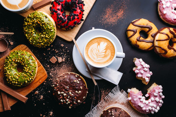Flat lay of various doughnuts with sweet toppings and chocolate bars composed with cup of cappuccino on black table