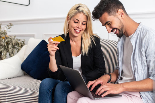 Delighted blonde female smiling and reading credit card credentials to cheerful ethnic boyfriend with laptop while sitting on sofa and making online purchases together