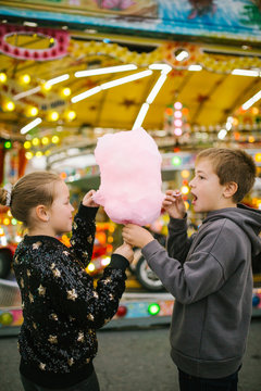 Side view of delighted girl and boy siblings smiling and eating sweet candyfloss while standing at funfair