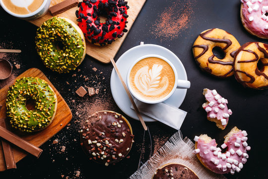 Flat lay of various doughnuts with sweet toppings and chocolate bars composed with cup of cappuccino on black table - Powered by Adobe