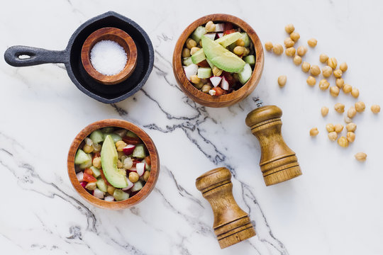 Flat lay of arranged wooden bowls with avocado salad with chickpeas composed on white marble table with salt and pepper seasonings