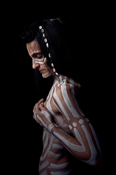 Side view of naked tender beautiful brunette Native American woman with white striped painted on body covering breast standing in dark on black background