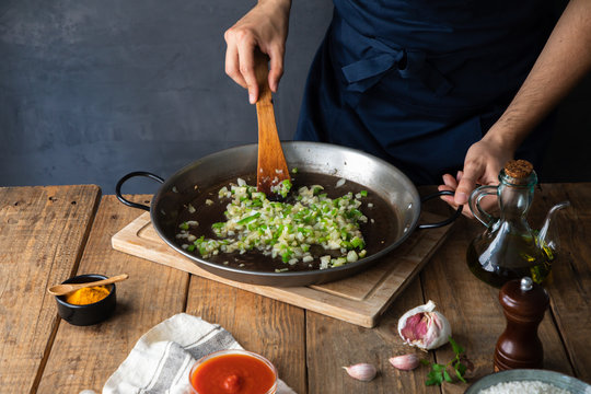 High angle of crop cook in dark apron stirring with wooden spatula mix of chopped green chili pepper and onion on big metal pan while adding oil before firing in contemporary kitchen at home