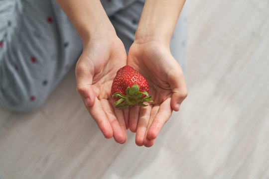 From above top view of anonymous kid sitting on floor and demonstrating fresh strawberry on summer day at home