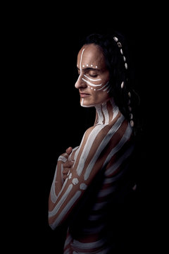 Side view of naked tender beautiful brunette Native American woman with white striped painted on body covering breast standing in dark on black background