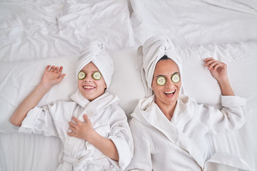 From above of happy young female and kid wearing white bathrobes and towel turbans lying on bed with cucumber slices on eyes and laughing while having fun and enjoying beauty procedure at home