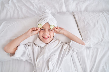Cheerful kid with slices of cucumber on yes lying on bed