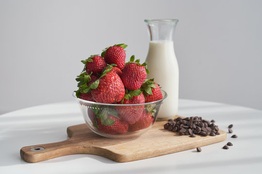 From above bowl of ripe strawberries and heap of chocolate pellets placed on napkin and cutting board near bottle of milk on kitchen table in morning
