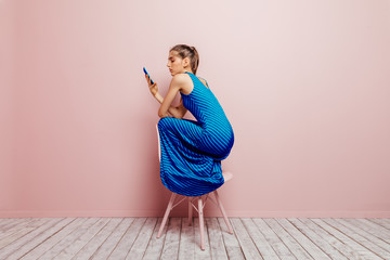 Side view of young woman crouched on top of a chair while using mobile phone on pink background