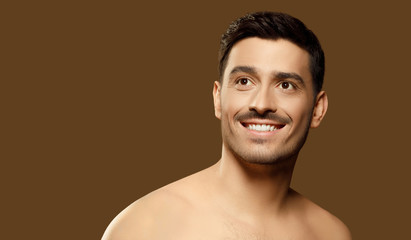 Horizontal banner of young male beauty model standng with naked shoulders and looking aside as if dreaming of something pleasant, skin care concept, isolated on brown background