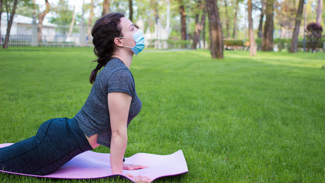 Young athletic woman in medical protective mask practicing yoga workout outdoor in park. Active life, sport during quarantine, coronavirus, covid-19. Concept of healthy lifestyle