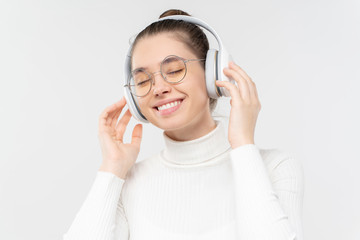 Closeup of young girl wearing glasses and wireless headphones, listening to music with eyes closed with pleasure, smiling, isoalted on gray background