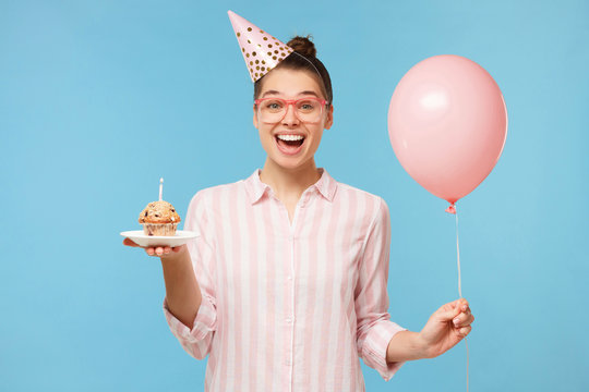 Young excited girl in birthday hat, holding pink balloon in one hand and festive cake in another, laughing happily, isolated on blue background