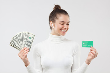 Young woman in white turtleneck, choosing between using dollars in cash or money on credit card, isolated on gray background