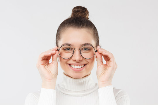 Close-up of smiling young nerdy girl, smart female student touching rim of round glasses as if trying to see something better, isolated on gray background