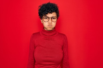Fototapeta na wymiar Young beautiful african american afro woman wearing turtleneck sweater and glasses puffing cheeks with funny face. Mouth inflated with air, crazy expression.
